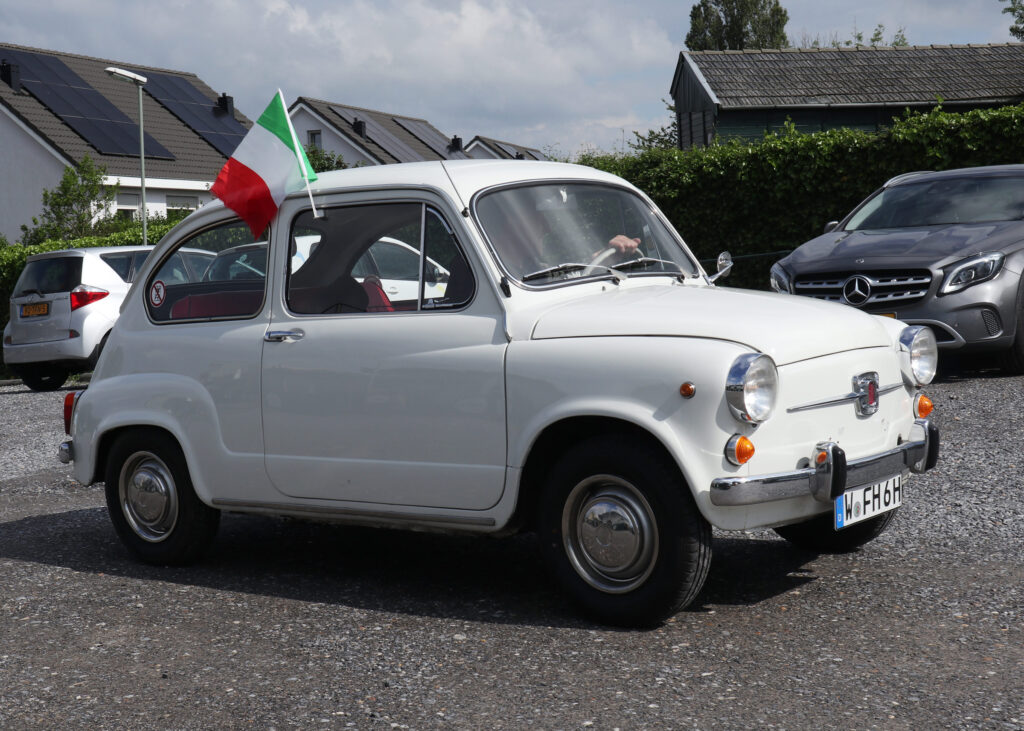 Fiat 600 mit italienischer Fahne am Seitenfenster fährt vom Parkplatz